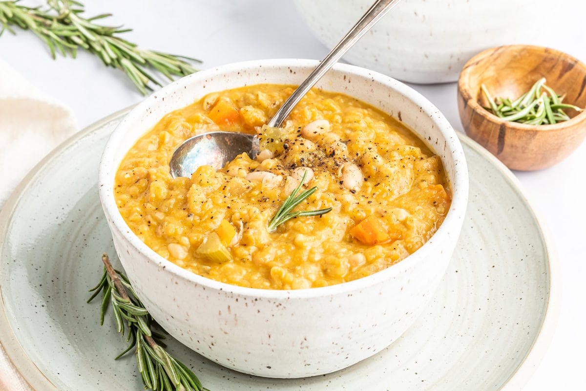 A bowl of creamy vegetable and bean stew garnished with rosemary, served with a spoon on a plate.