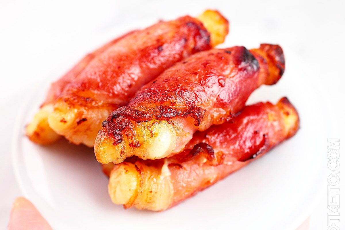 Three bacon-wrapped cheese sticks stacked on a white plate.