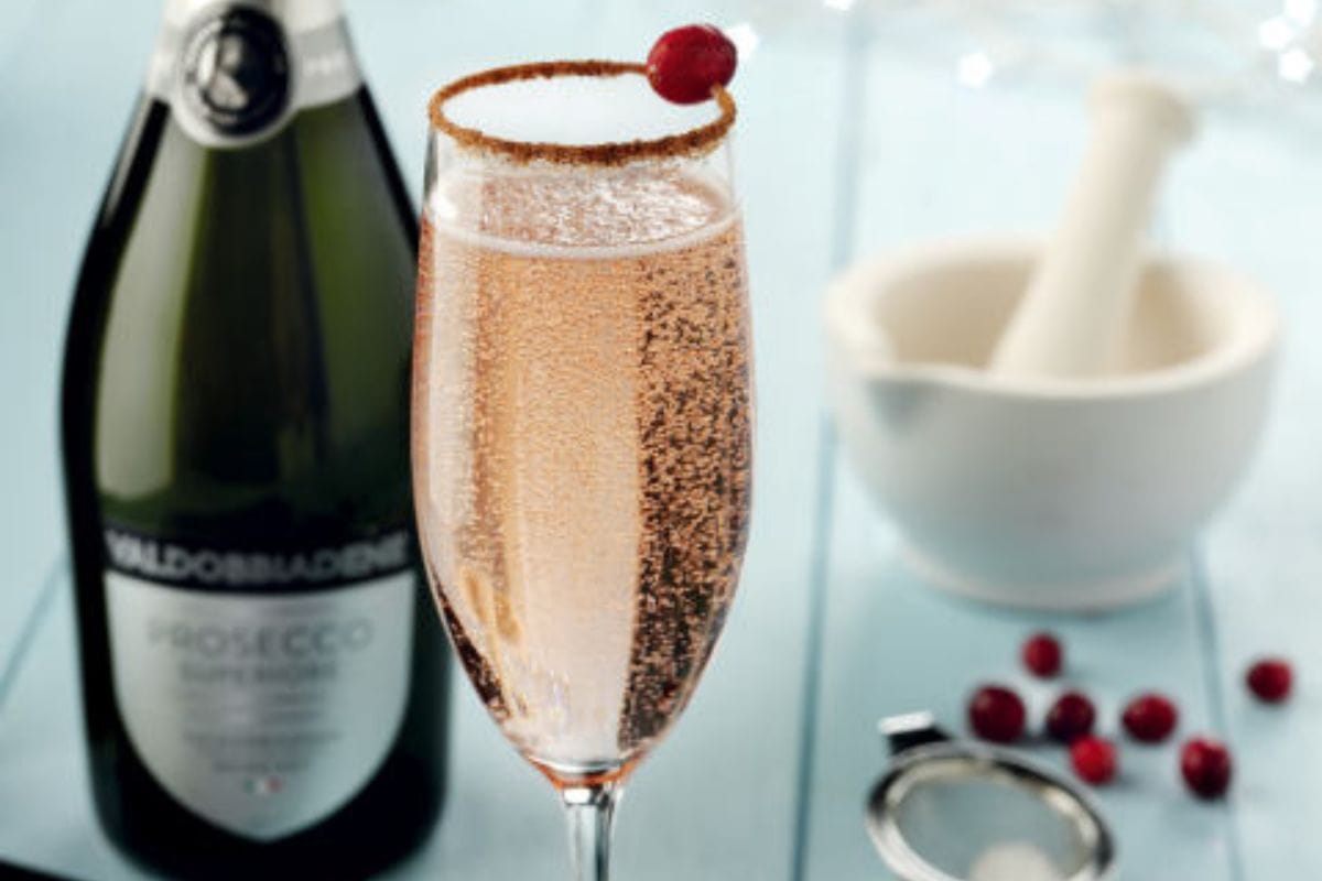 A glass of pink Prosecco with a sugared rim and cranberry garnish, perfect for easy holiday cocktails, sits beside a bottle of the sparkling delight. In the background, a mortar and pestle teams up with scattered cranberries to complete the festive scene.