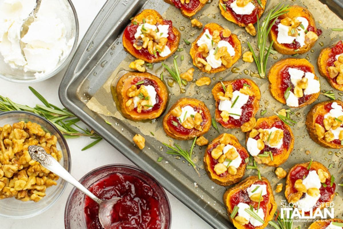 Sweet potato rounds topped with cranberry sauce, walnuts, and cheese on a baking sheet, next to bowls of cream, walnuts, and cranberry sauce. Garnished with rosemary.