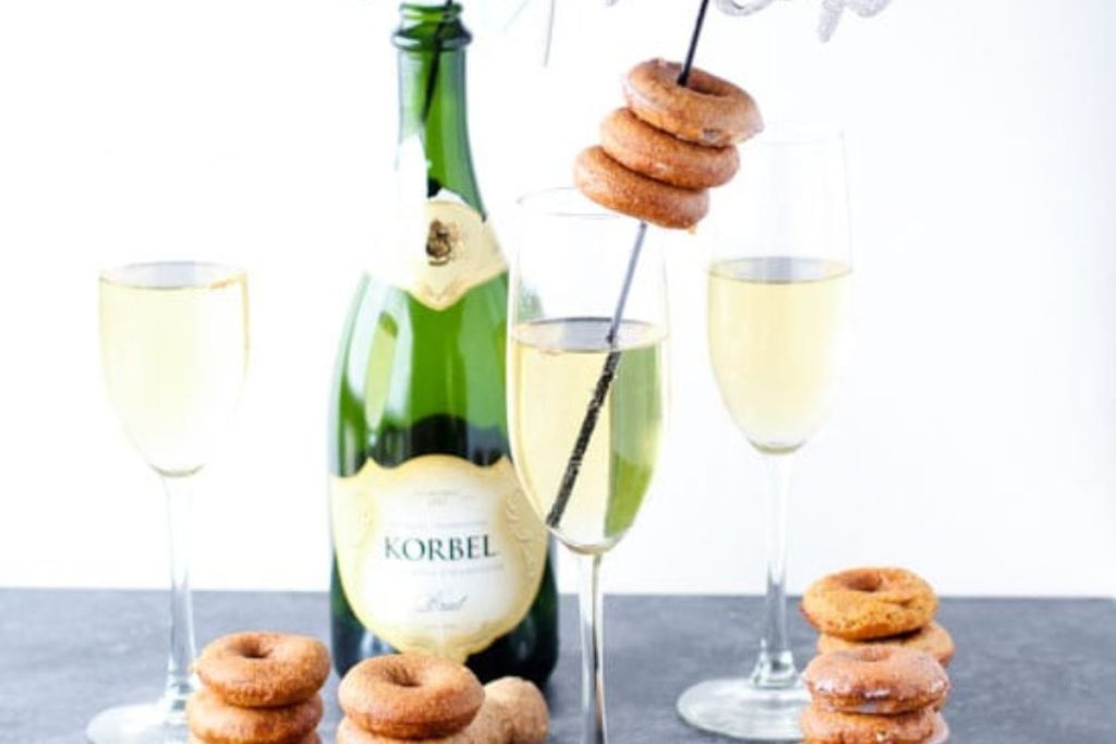 A bottle of Korbel champagne with two filled flutes, perfect for New Year's Eve. Mini donuts are stacked on a black straw resting over one glass, adding a sweet twist to your appetizers. Additional mini donuts are scattered around for festive snacking.