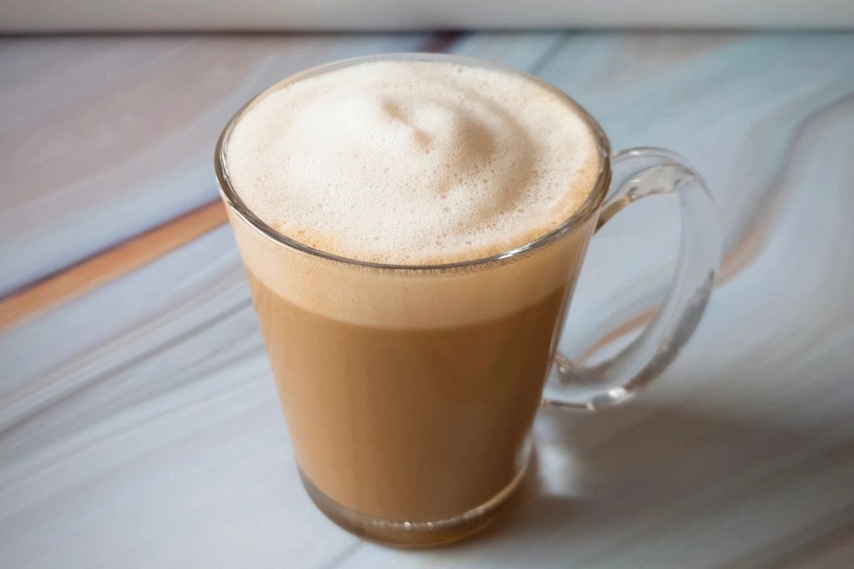 A glass mug filled with frothy coffee sits on a marbled surface.