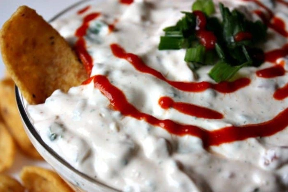 A bowl of creamy holiday dip topped with a drizzle of red sauce and garnished with green onions. A chip is dipped into the mixture.