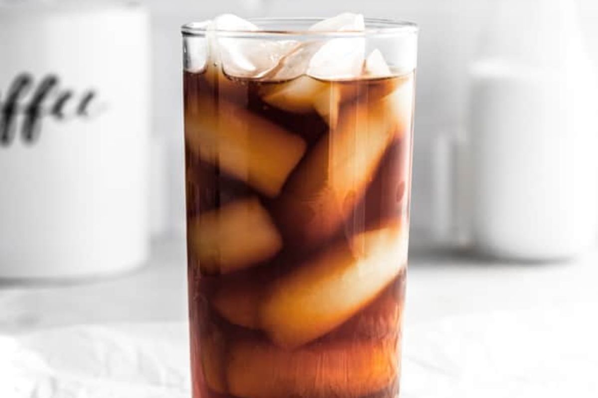 A glass of iced coffee with ice cubes on a table, blurry background featuring a coffee container and a milk jug.