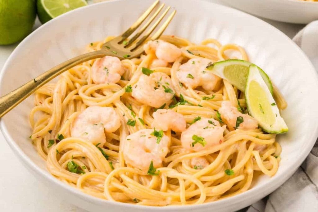 A bowl of spaghetti with shrimp, garnished with lime slices and herbs, offers a delightful twist for your soul food dinner. A fork rests on the edge of the bowl, inviting guests to indulge in this delicious menu idea.