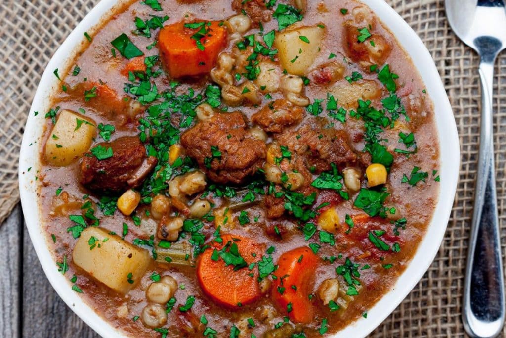A bowl of hearty beef stew with carrots, potatoes, peas, and parsley is the perfect soul food dinner. It's placed on a textured mat with a spoon and knife beside it.