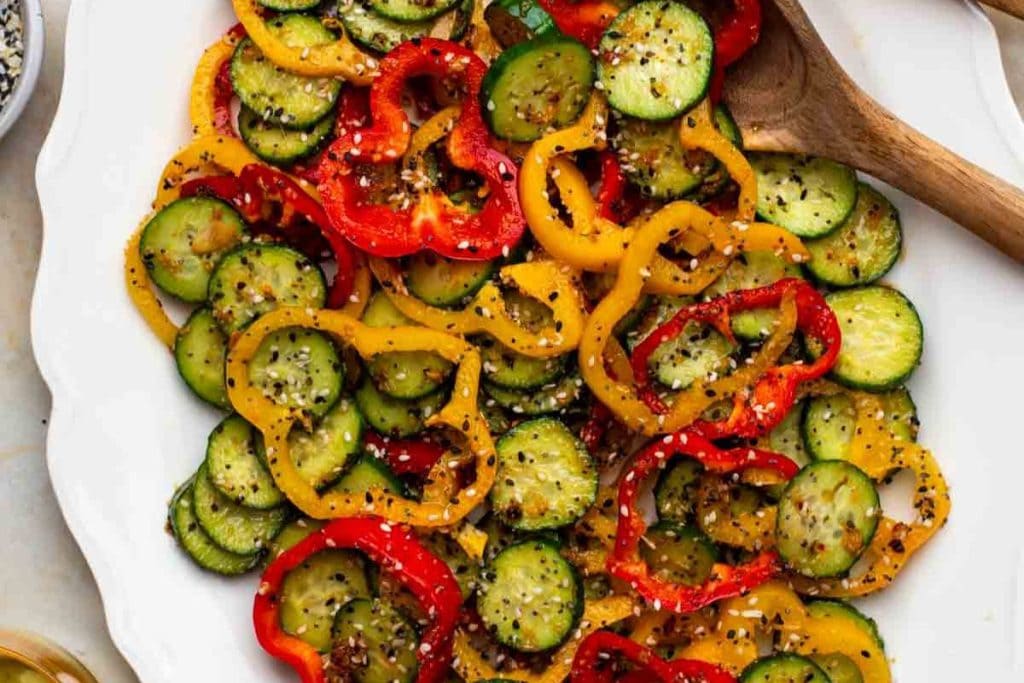 A plate of sliced cucumbers and red and yellow bell peppers, sprinkled with seasoning, showcases one of the simplest yet most delightful healthy salad recipes. Accompanied by a wooden spoon, it invites you to dig into its fresh crunchiness.