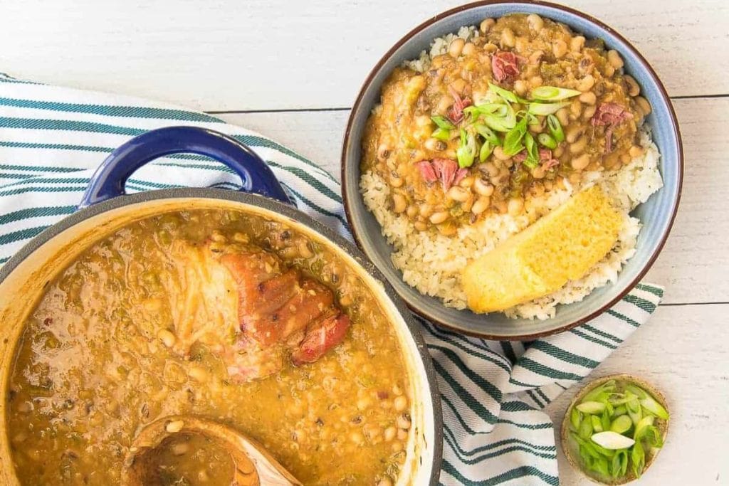 A soul food dinner featuring a pot of stew with beans and meat sits beside a bowl of rice topped with the rich stew, cornbread, and green onions. A striped towel and a small bowl of fresh green onions add charm to this comforting meal.