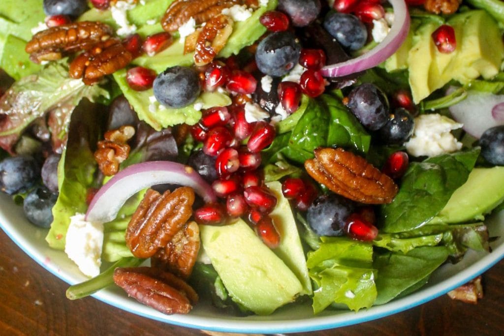 A healthy salad recipe featuring mixed greens, creamy avocado slices, crunchy pecans, juicy blueberries, vibrant pomegranate seeds, tangy red onion slices, and crumbled cheese all tossed in a bowl.