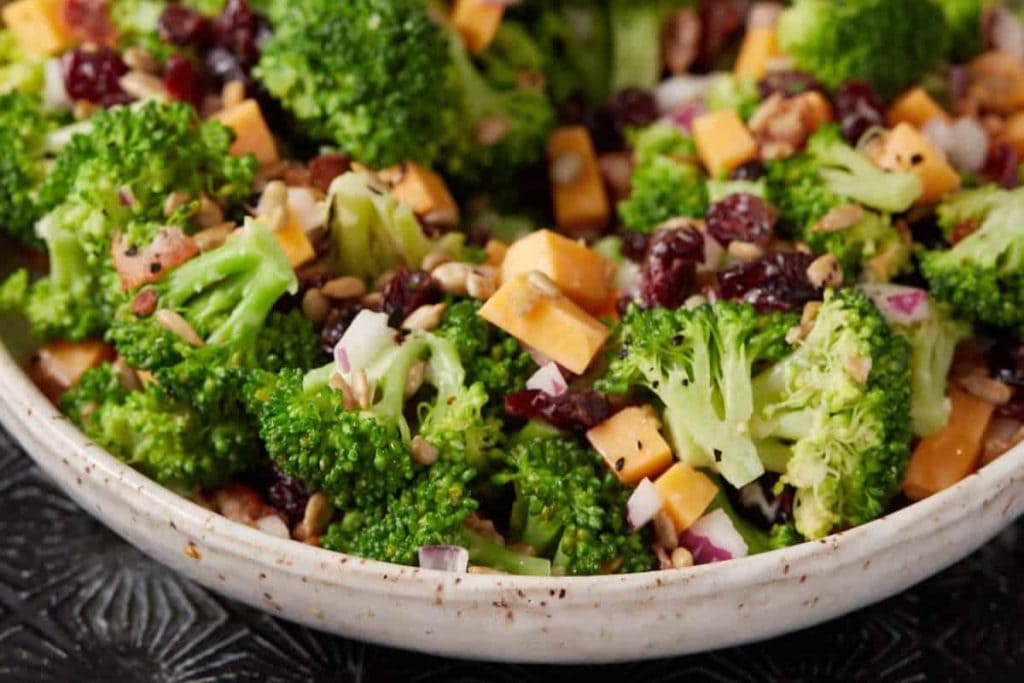 A bowl of broccoli salad, featuring cheddar cheese cubes, dried cranberries, sunflower seeds, and chopped red onion, showcases one of the delightful healthy salad recipes on a textured surface.