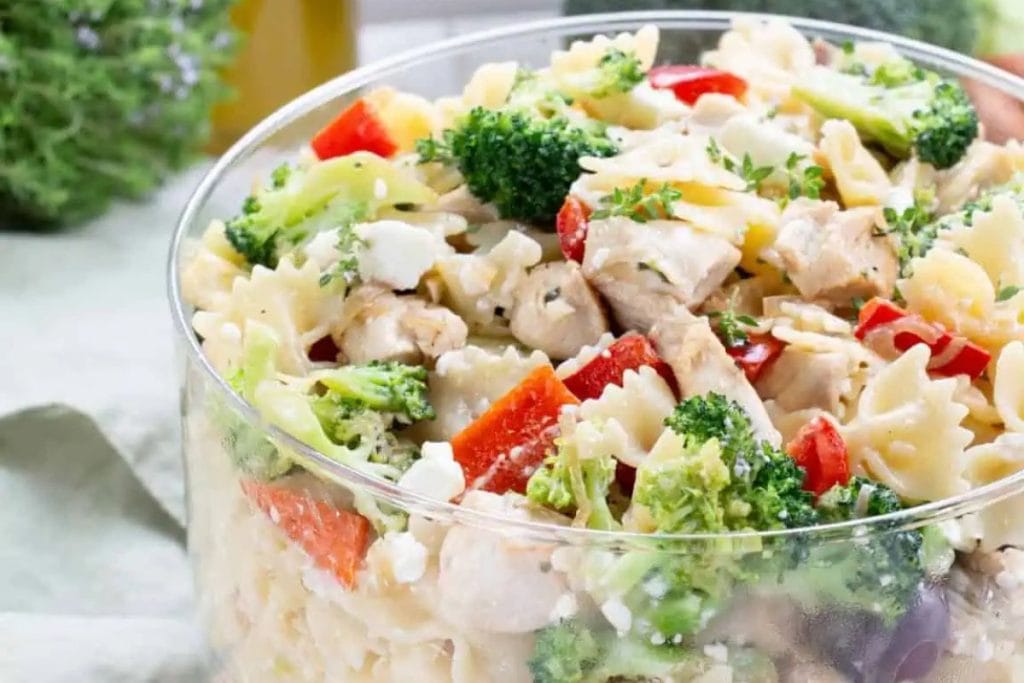 A glass bowl brimming with pasta salad—featuring bowtie pasta, broccoli, red bell peppers, chicken, and herbs—is poised on a table with a blurred background, making it an ideal on-the-go lunch.