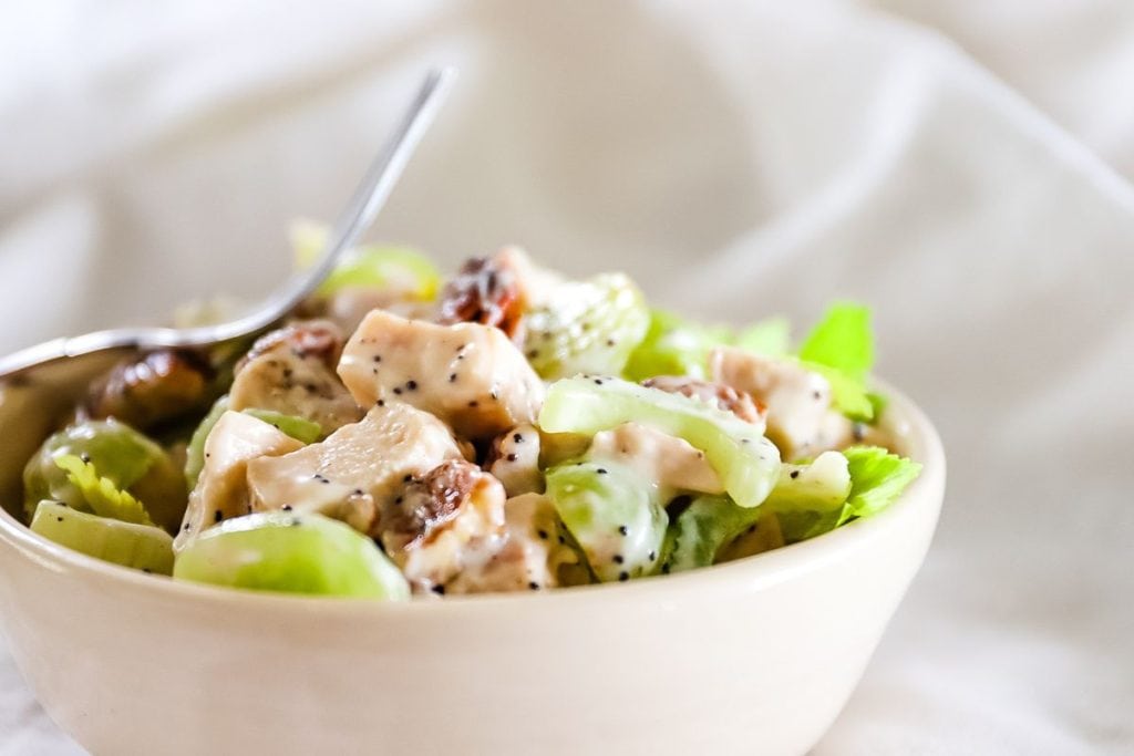 A bowl of chicken salad featuring crisp celery and juicy grapes, drizzled with a light dressing, rests invitingly on a white cloth with a spoon poised to serve. Perfect for those seeking healthy chicken recipes that burst with fresh flavors.