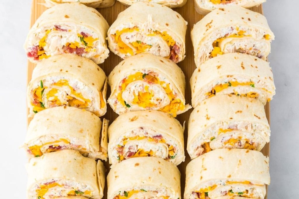 Assorted tortilla pinwheels filled with sliced turkey, cheese, lettuce, and tomato, arranged in rows on a wooden surface.