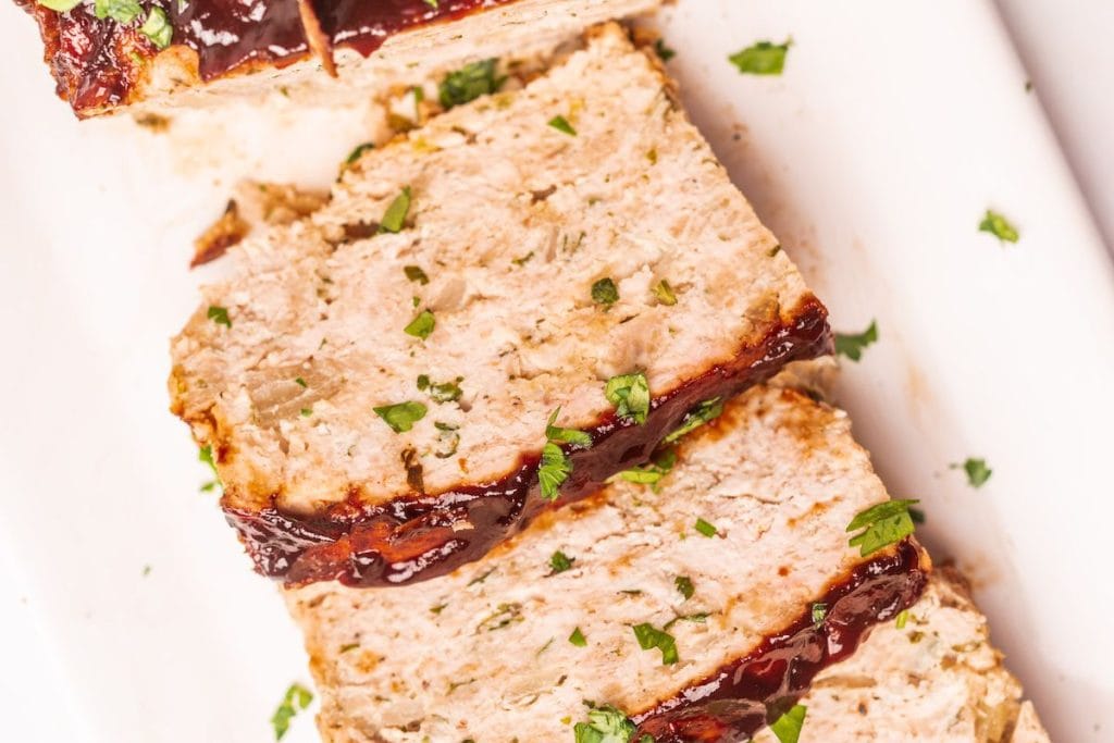 Slices of healthy, chicken-inspired meatloaf topped with sauce and garnished with chopped parsley on a white plate.
