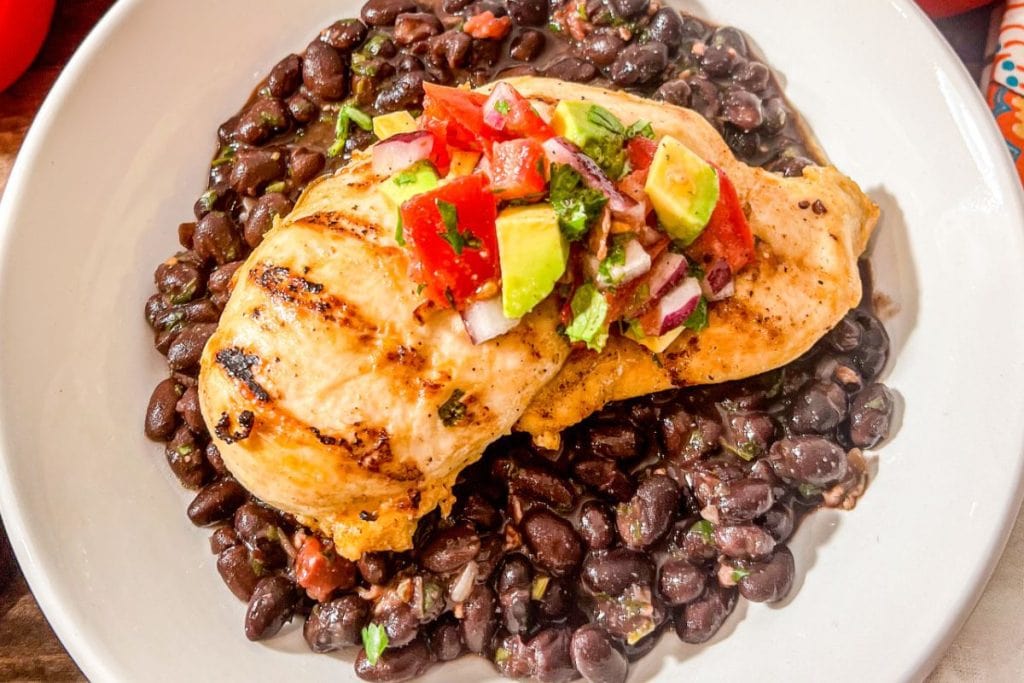 Grilled chicken breast, a staple in healthy chicken recipes, is topped with vibrant salsa and rests invitingly on a bed of black beans on a white plate.