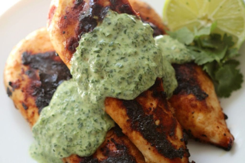 Grilled chicken breast with a charred surface, topped with green herb sauce, garnished with lime slices and cilantro on the side. This dish is a perfect addition to any collection of healthy chicken recipes.