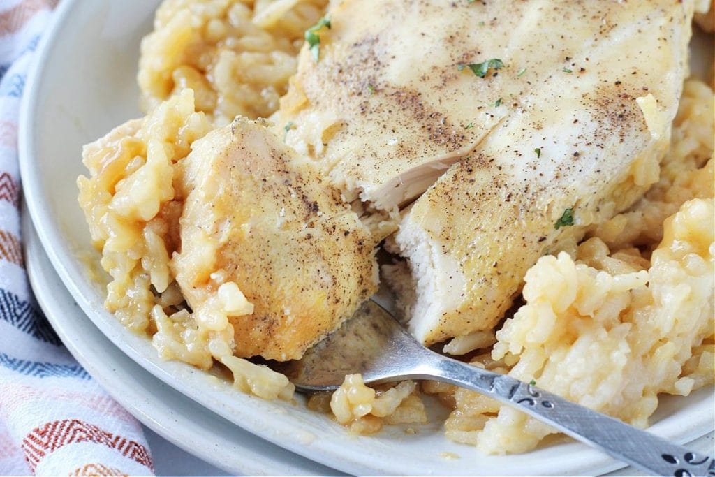 A fork cutting into seasoned baked chicken on a plate, served with creamy rice—a perfect addition to your soul food dinner party menu.