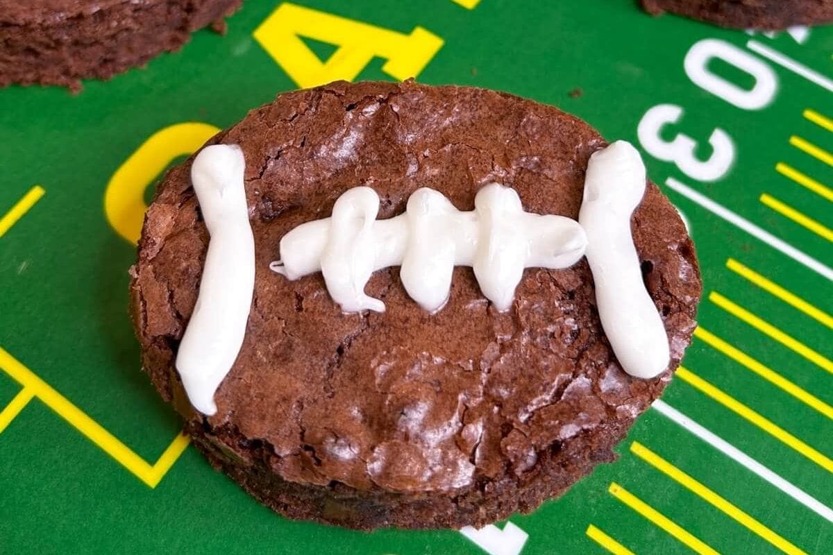 A football dessert masterpiece, this brownie is artfully decorated to resemble a football, complete with white frosting laces. It's set on a green surface adorned with football field markings, making it a perfect match for game day celebrations.