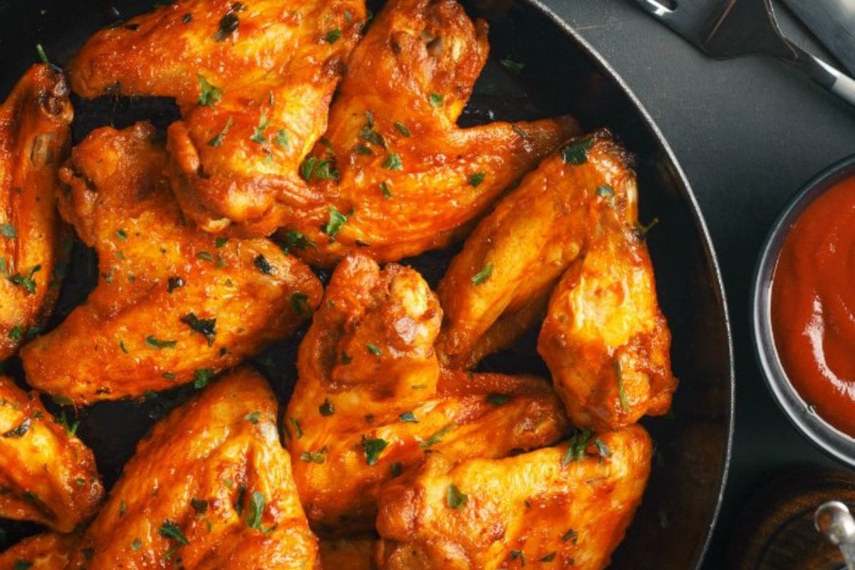 A pan of seasoned, cooked chicken wings garnished with herbs makes the perfect football party appetizer. A small bowl of red sauce is ready on the side for dipping.