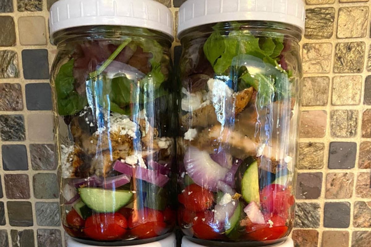 Two jars filled with a layered salad, including cherry tomatoes, cucumbers, red onions, feta cheese, greens, and grilled chicken slices stand against a tiled backsplash—perfect meal prep ideas for a healthy week ahead.