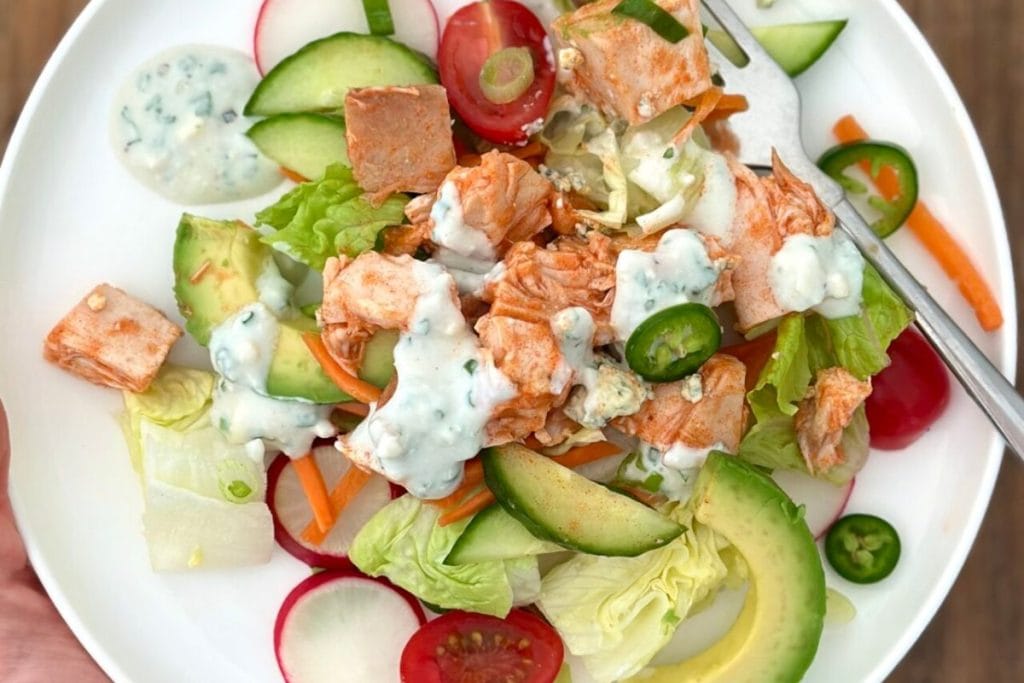 A plate of healthy salad features lettuce, cherry tomatoes, cucumber, sliced radishes, avocado, shredded carrots, diced chicken, jalapeño slices, and a dollop of creamy dressing.