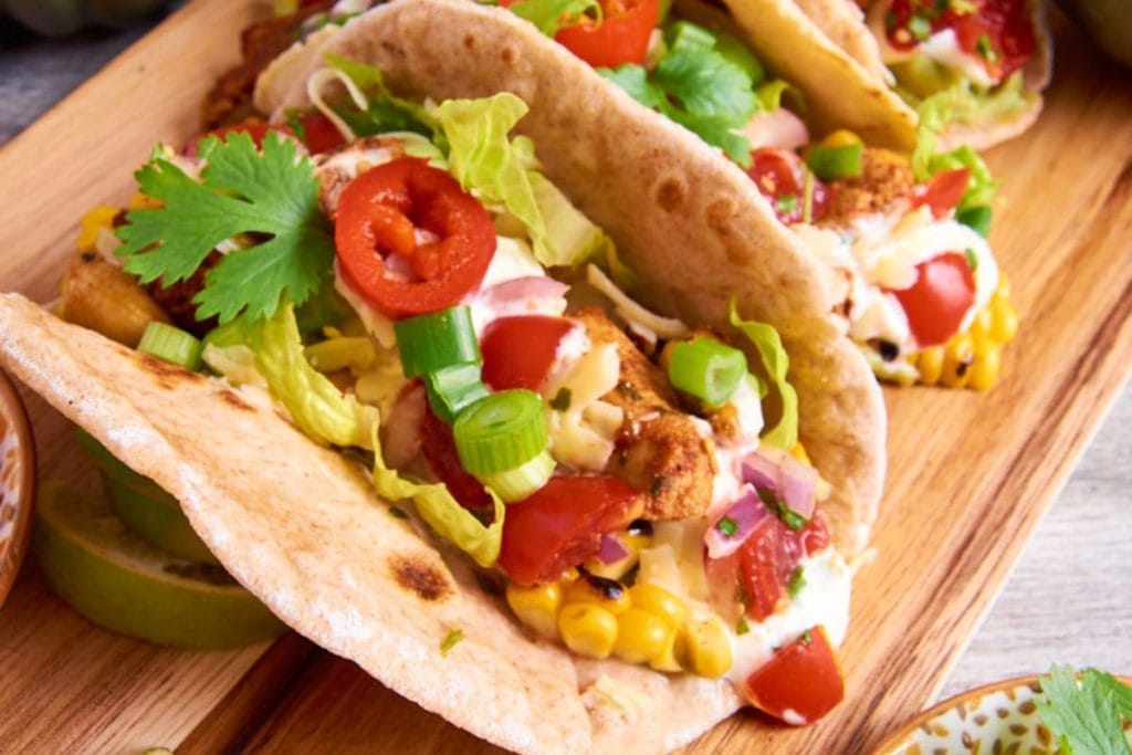 Close-up of colorful tacos filled with lettuce, tomatoes, corn, green onions, and sliced red peppers on a wooden board. These vibrant delights are a perfect addition to your list of healthy chicken recipes.