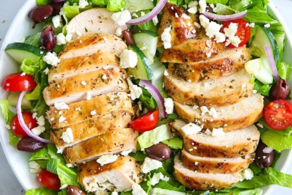 A fresh and healthy salad recipe featuring sliced grilled chicken, feta cheese, cherry tomatoes, cucumber, olives, and red onion on a bed of leafy greens.