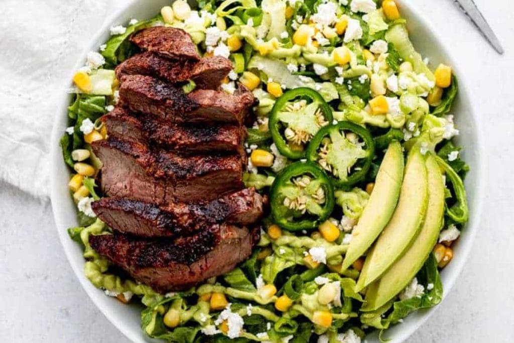 A healthy salad recipe featuring grilled steak, sliced jalapeños, avocado, corn, lettuce, and crumbled cheese served in a white bowl.