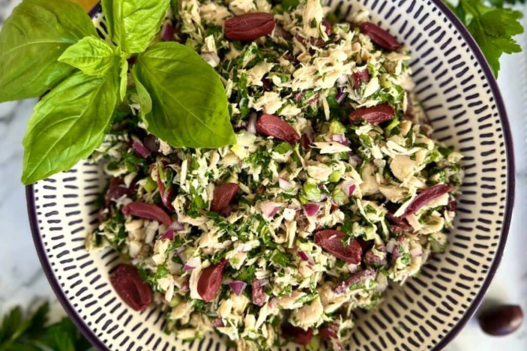 A bowl of tuna salad with chopped greens, red onions, and olives, garnished with fresh basil leaves.