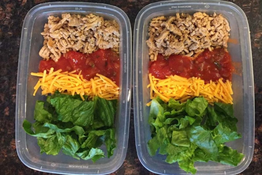 Two meal prep containers each with cooked ground meat, diced tomatoes, shredded cheddar cheese, and leafy greens, arranged in sections.