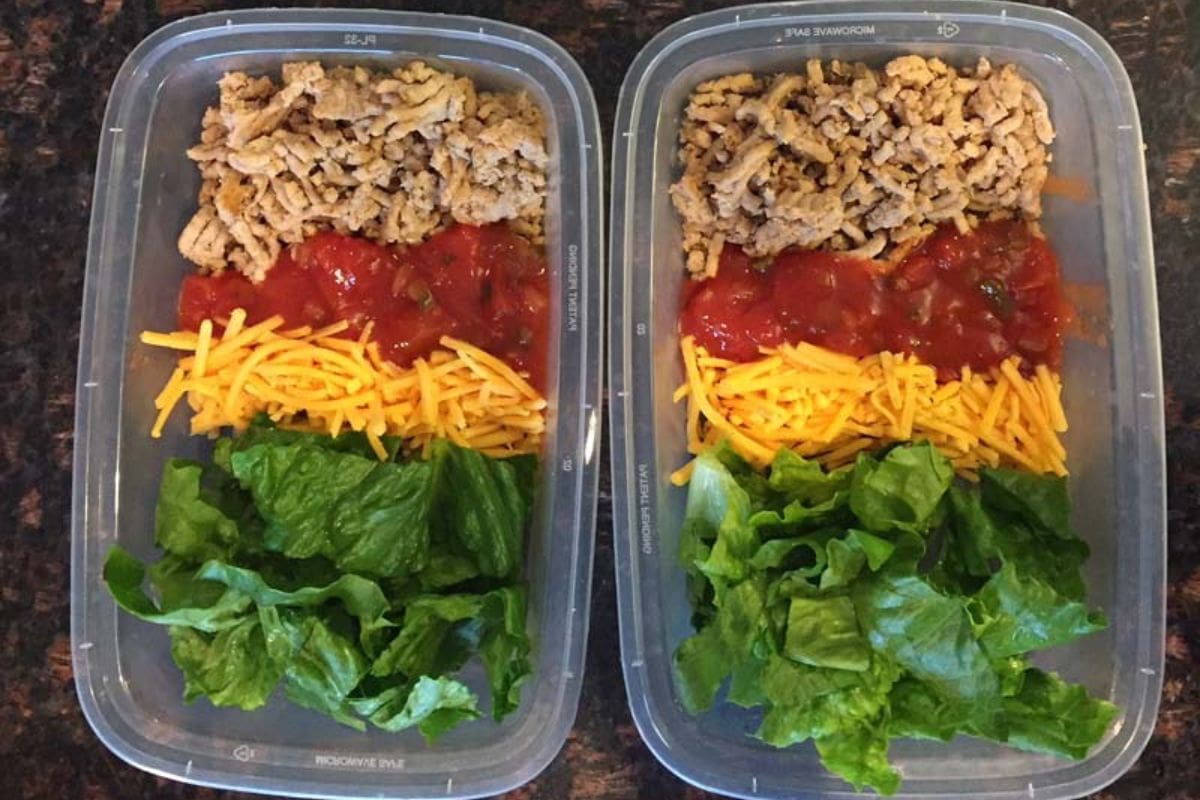 Two meal prep containers each with cooked ground meat, diced tomatoes, shredded cheddar cheese, and leafy greens, arranged in sections.