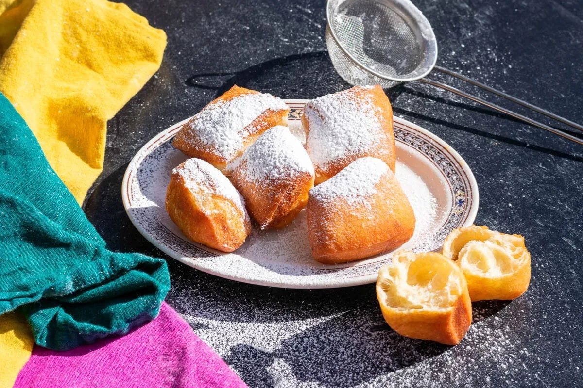 A plate with four beignets dusted with powdered sugar sits enticingly, exuding the festive spirit of Mardi Gras food. One beignet is halved, revealing its fluffy interior. A metal sieve and colorful cloths add to the vibrant scene.