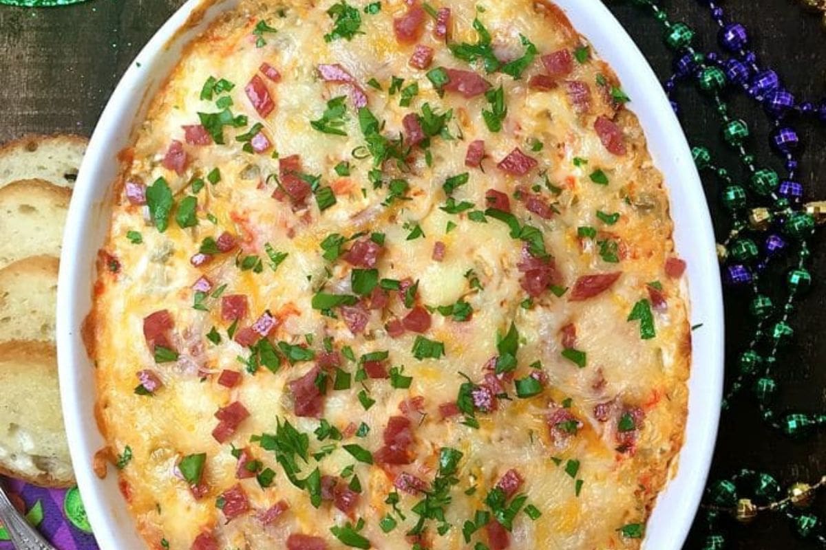 Oval dish with baked cheese dip topped with diced meat and fresh parsley, surrounded by slices of bread and colorful beads on a dark surface, capturing the festive spirit of Mardi Gras food.