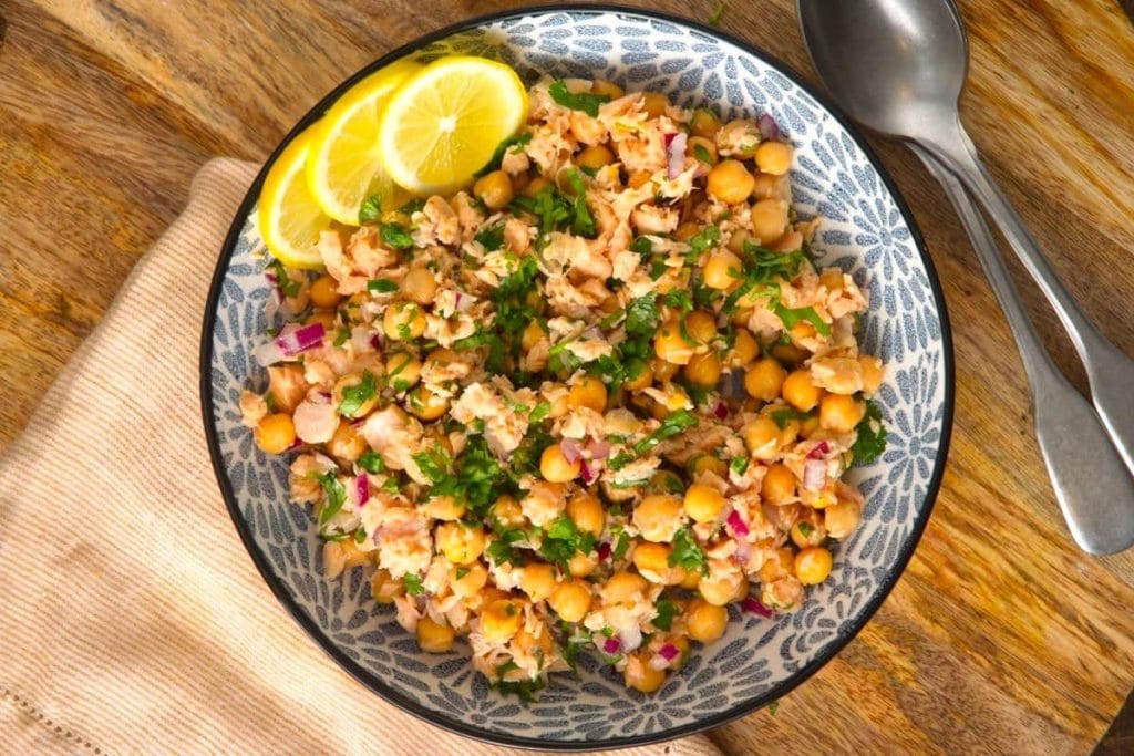 A bowl of chickpea salad with parsley, onions, and shredded chicken, garnished with lemon slices. A fork and spoon are placed beside the bowl on a wooden surface.