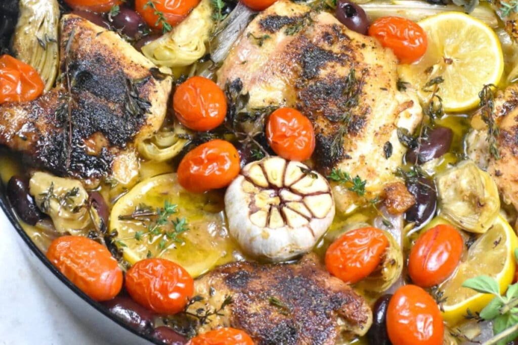 Enjoy this healthy chicken recipe featuring roasted chicken thighs with cherry tomatoes, artichokes, olives, lemon slices, and a whole garlic bulb in a pan.