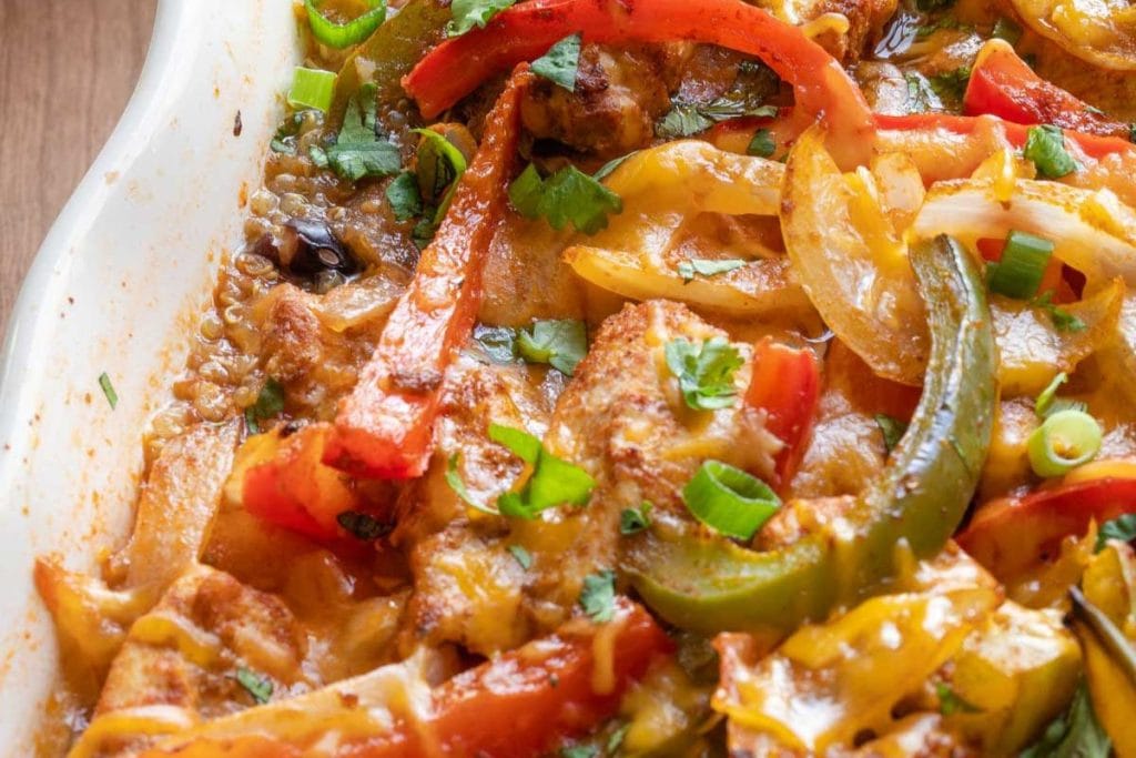 Close-up of a baked chicken fajita dish with sliced red, yellow, and green bell peppers, topped with melted cheese and chopped green onions in a white baking dish.