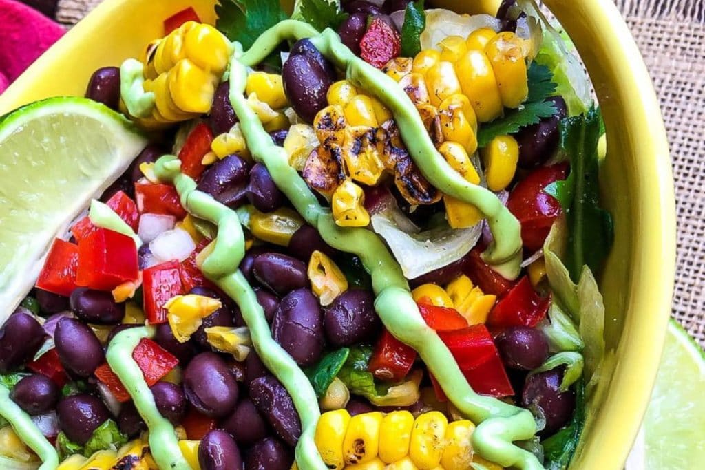 A bowl of healthy salad featuring grilled corn, black beans, red bell peppers, and avocado dressing, garnished with lime wedges.
