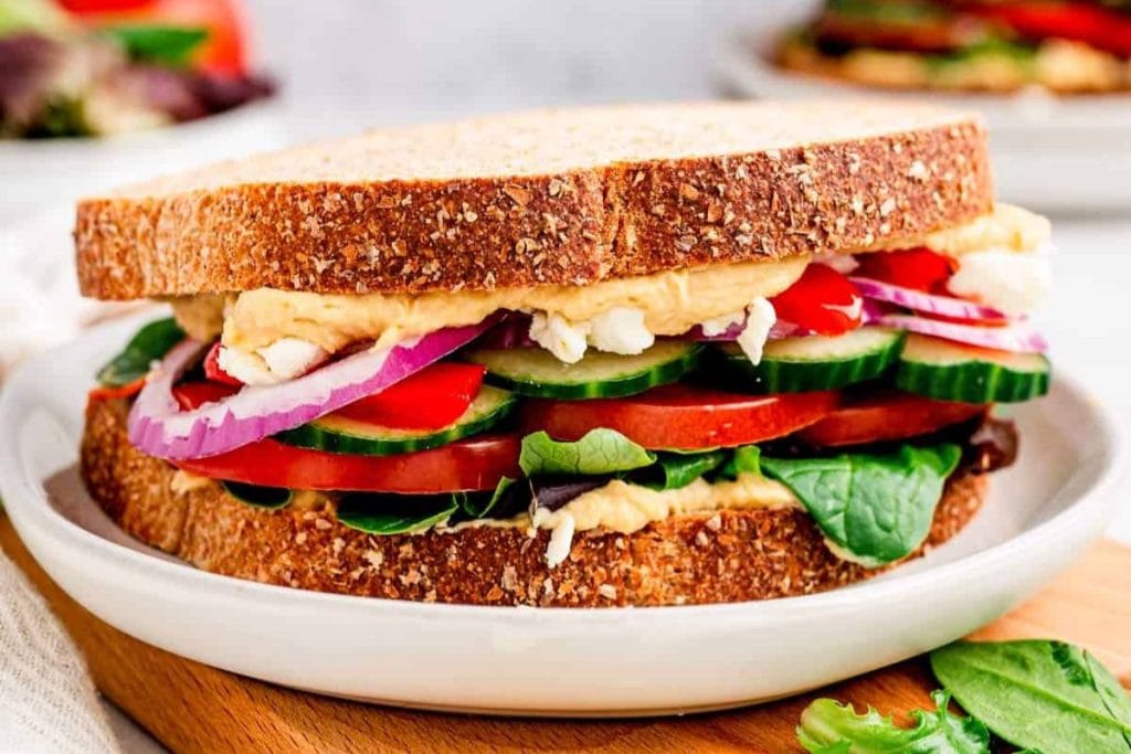 A close-up of a sandwich with multigrain bread, perfect for an on-the-go lunch, layered with hummus, red onion, cucumber, tomato, and greens on a white plate.