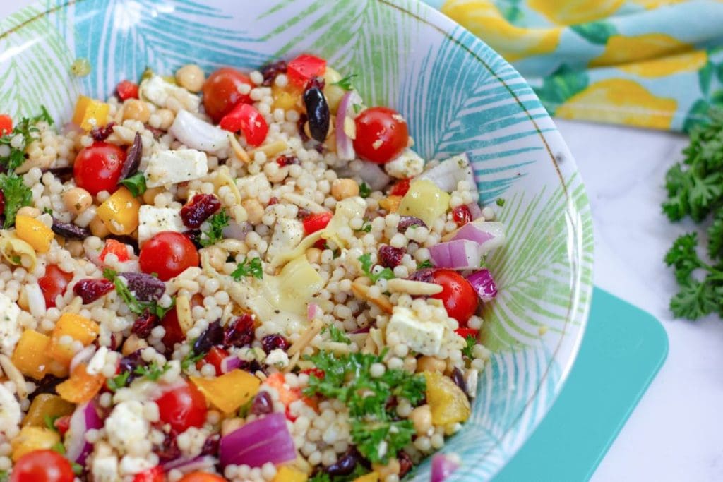 A colorful couscous salad, perfect for healthy salad recipes, features cherry tomatoes, chickpeas, feta, red onion, olives, and fresh herbs artfully arranged in a patterned bowl.