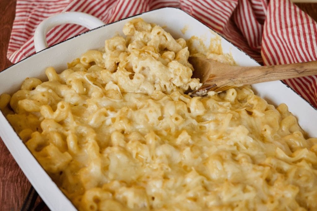 A white baking dish brimming with creamy baked macaroni and cheese, exuding the comforting essence of a soul food dinner. A wooden spoon rests atop it, while a red and white striped cloth peeks from the background.