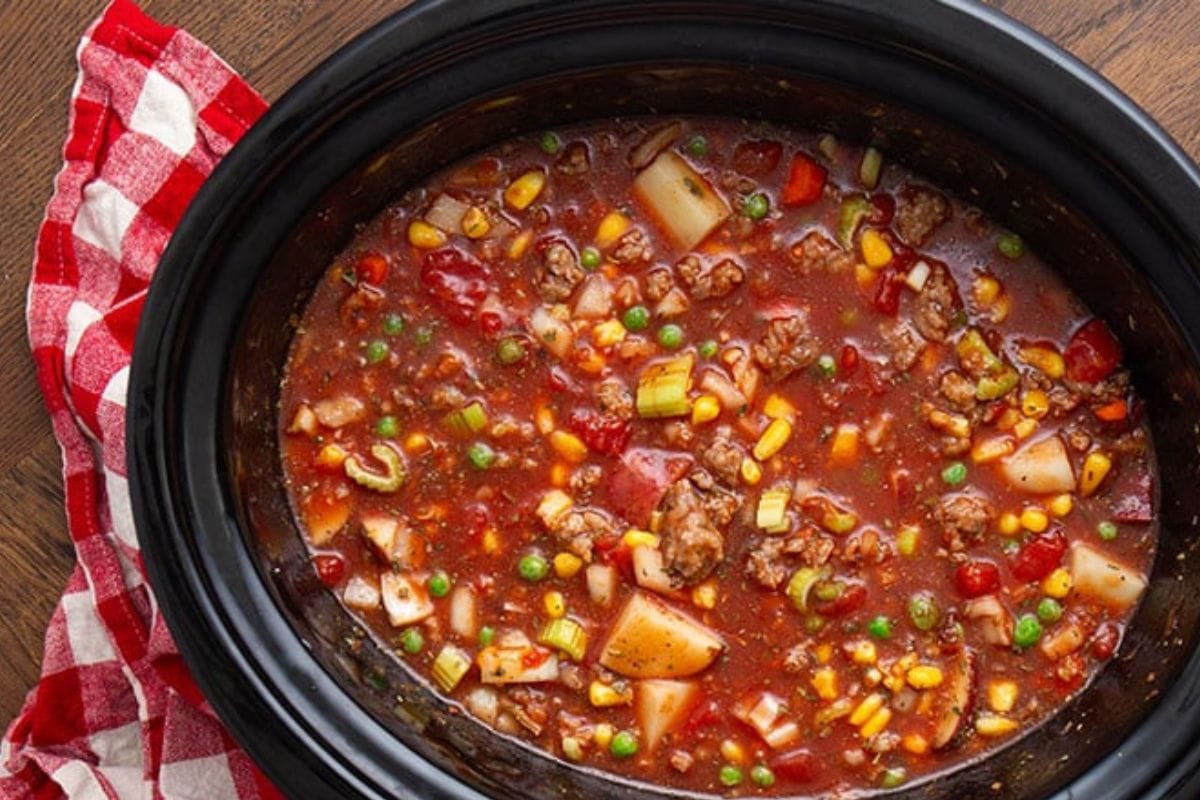 A slow cooker filled with vegetable and meat stew, including corn, peas, potatoes, and peppers, next to a red and white checkered cloth.