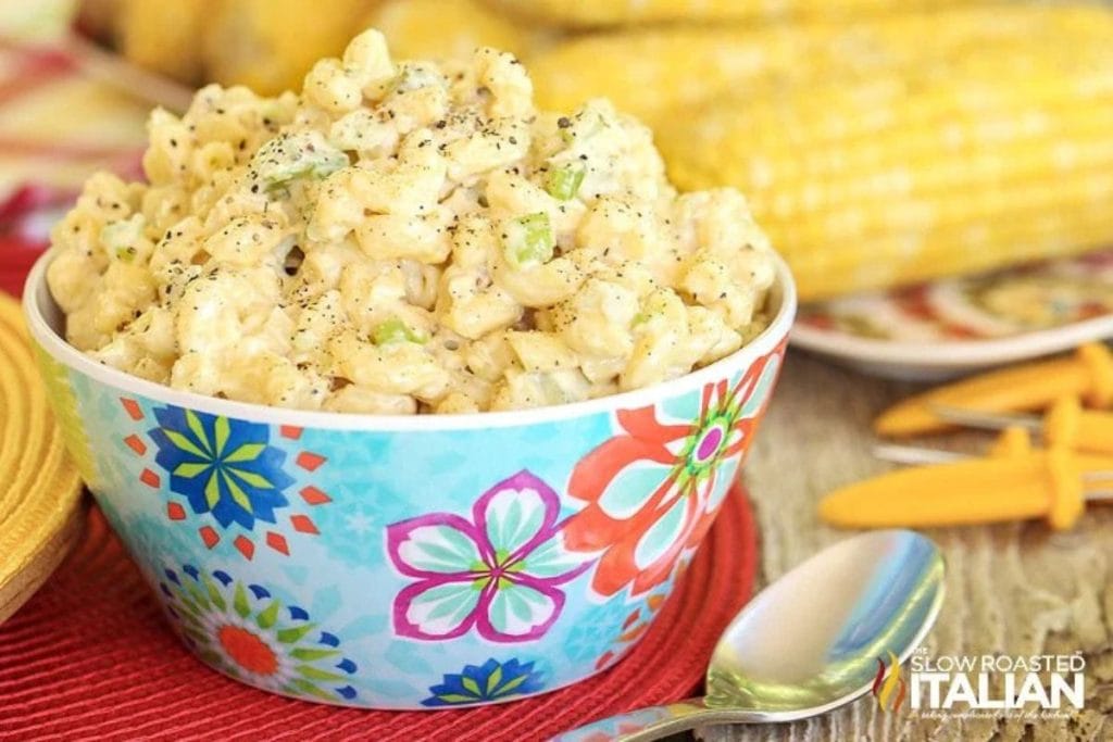 A colorful bowl of creamy macaroni salad, garnished with pepper, captures the essence of soul food. Perfect for inspiring dinner party menu ideas, corn on the cob and utensils linger in the background.