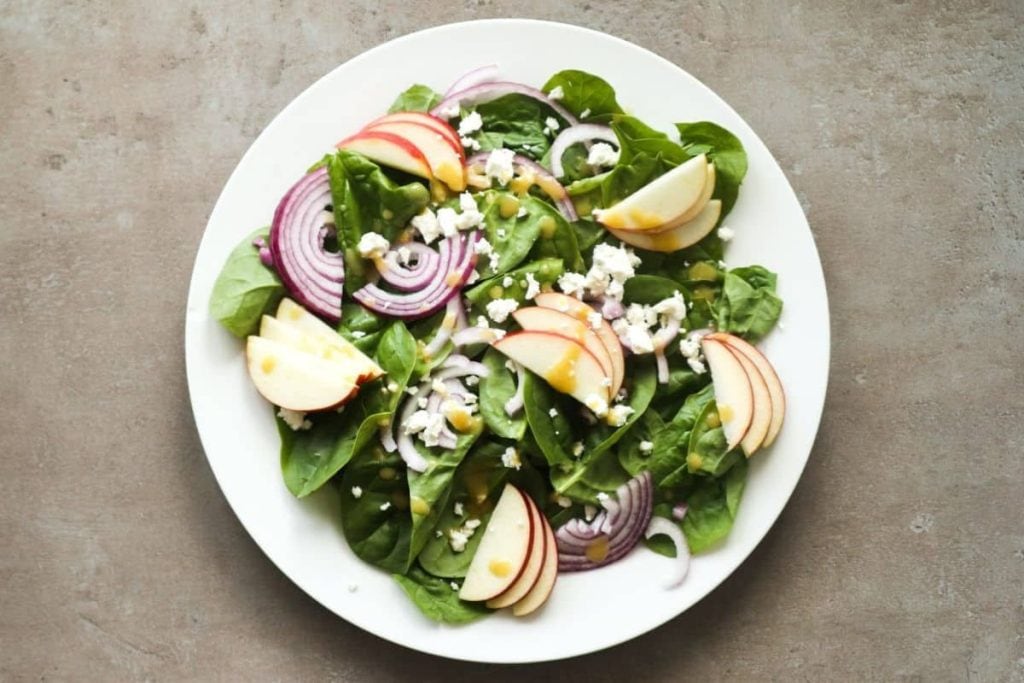 A healthy salad recipe features spinach, sliced apples, red onion, and creamy feta cheese on a white plate, all drizzled with a light dressing.