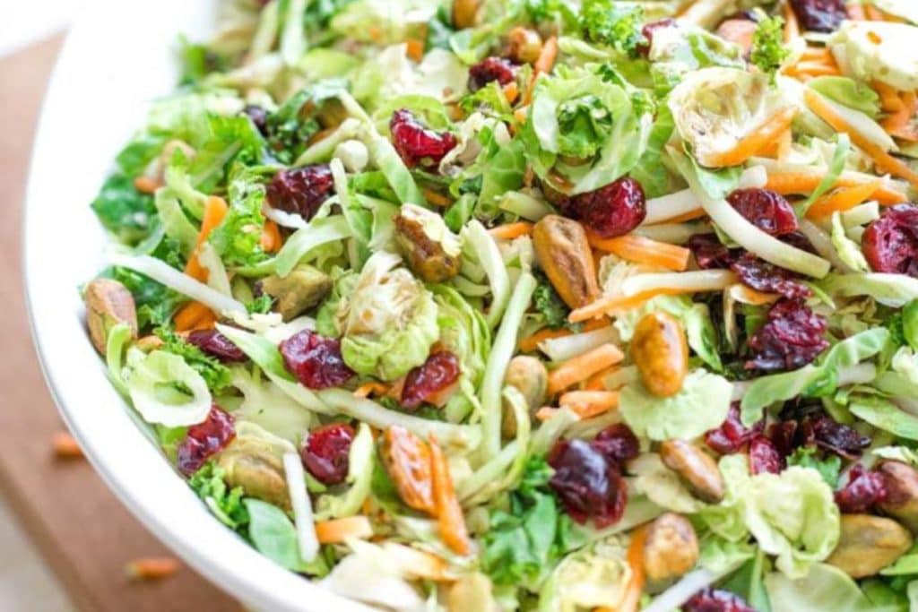 Discover healthy salad recipes with this vibrant dish: a salad in a white bowl featuring Brussels sprout leaves, shredded carrots, dried cranberries, and pistachios.