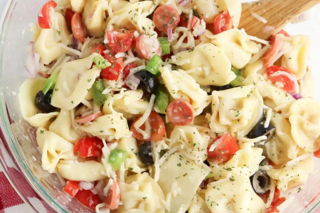 Bowl of tortellini salad with tomatoes, olives, green peppers, onions, and shredded cheese, mixed with a wooden spoon.