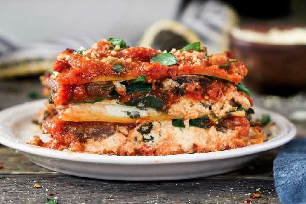 A slice of vegetable lasagna on a white plate, layered with spinach, ricotta, and tomato sauce, embodies a comforting soul food dinner. It's topped with fresh basil and grated cheese, creating a warm and inviting dish perfect for any evening.