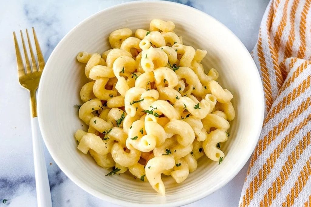A bowl of cooked cavatappi pasta topped with herbs sits ready for an on-the-go lunch, next to a fork on a marble surface and a striped cloth.