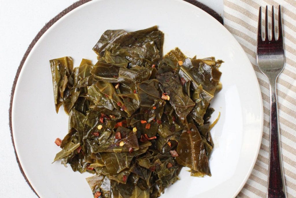 A plate of soul food featuring cooked collard greens sprinkled with red pepper flakes sits next to a fork on a striped napkin, offering perfect dinner party menu ideas.