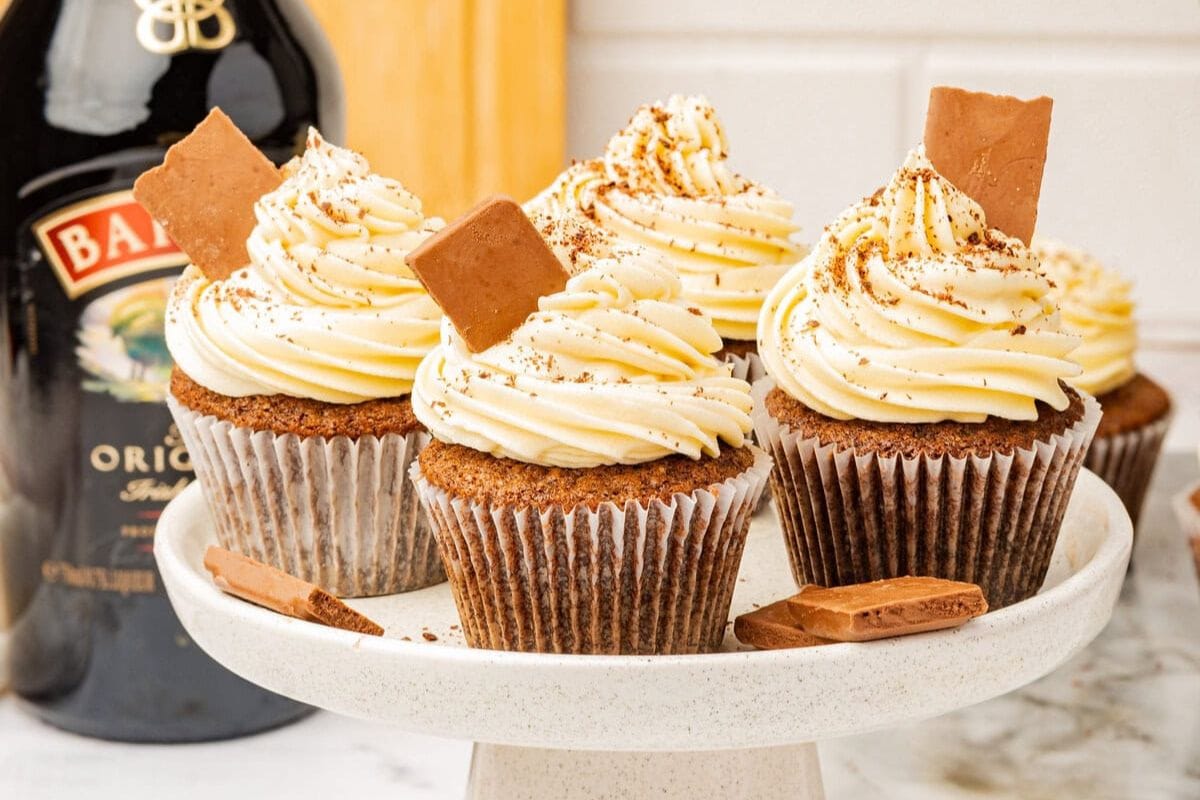 Cupcakes with swirled cream frosting are topped with chocolate pieces and displayed on a marble stand. A bottle is visible in the background.
