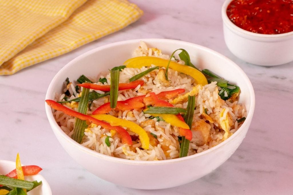 A bowl of fried rice with sliced red and yellow bell peppers, green onions, and egg on a marble surface. A small bowl of red sauce and a yellow cloth are in the background.
